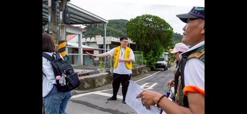 淡江大學志工手持指揮棒，在金崙部落路口引導健走隊伍前行，前方可見穿著傳統服飾的在地部落耆老共同參與。